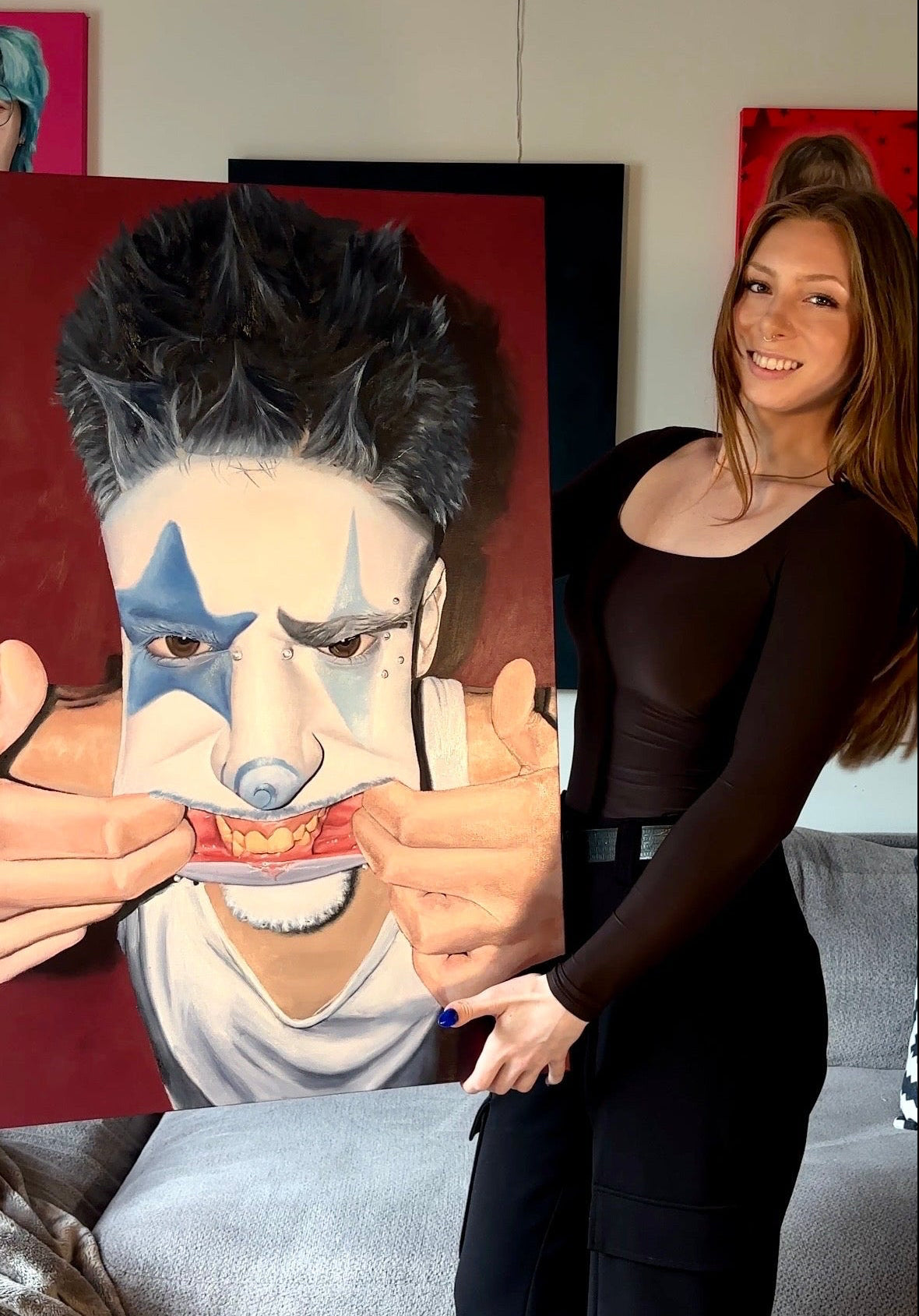 A woman stands indoors next to a large canvas painting of a person with blue face paint and black hair against a red background. The room includes a gray sofa, framed wall art, and a pink neon sign.