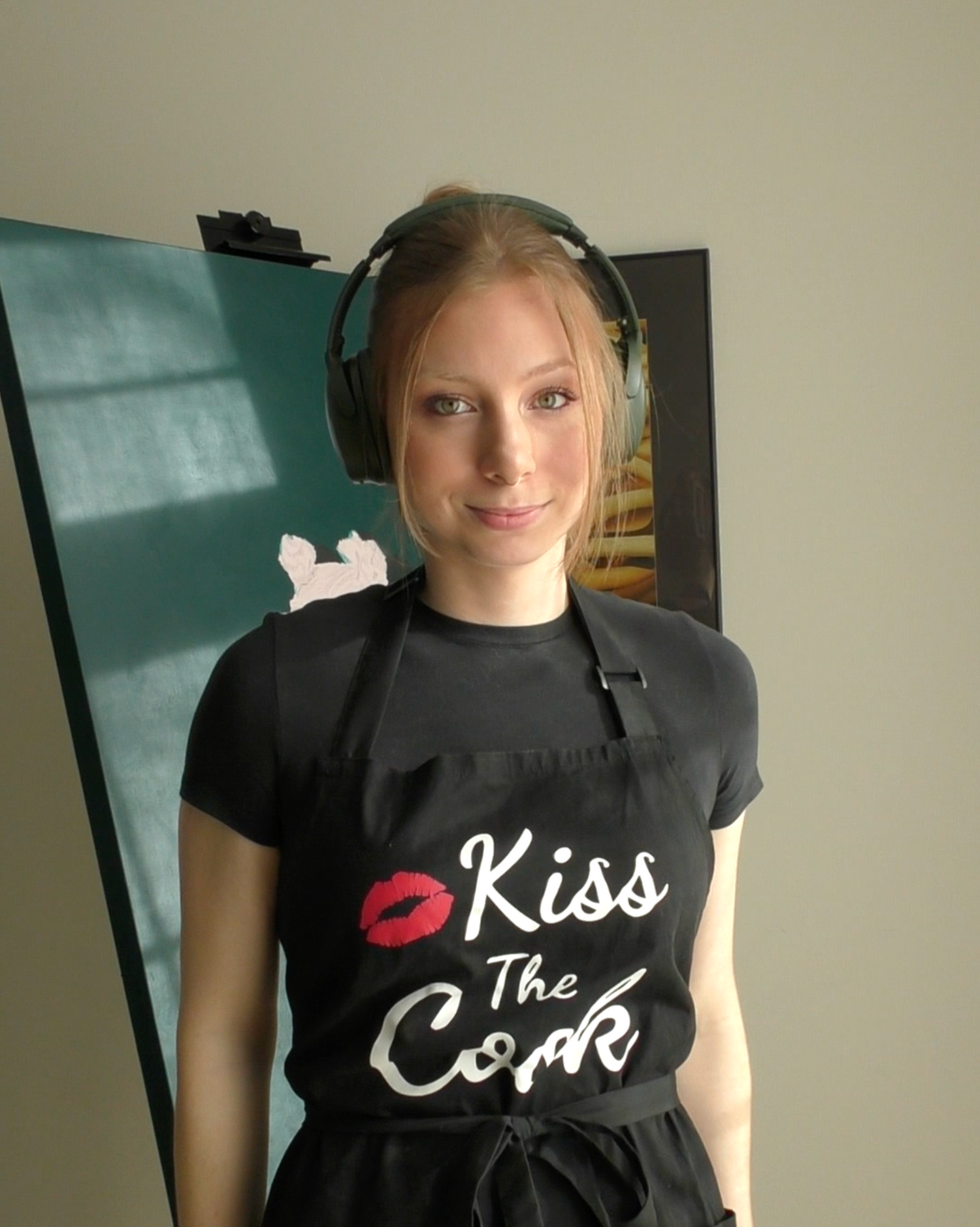 The artist wearing a black apron with 'Kiss The Cook' text and red lipstick design.