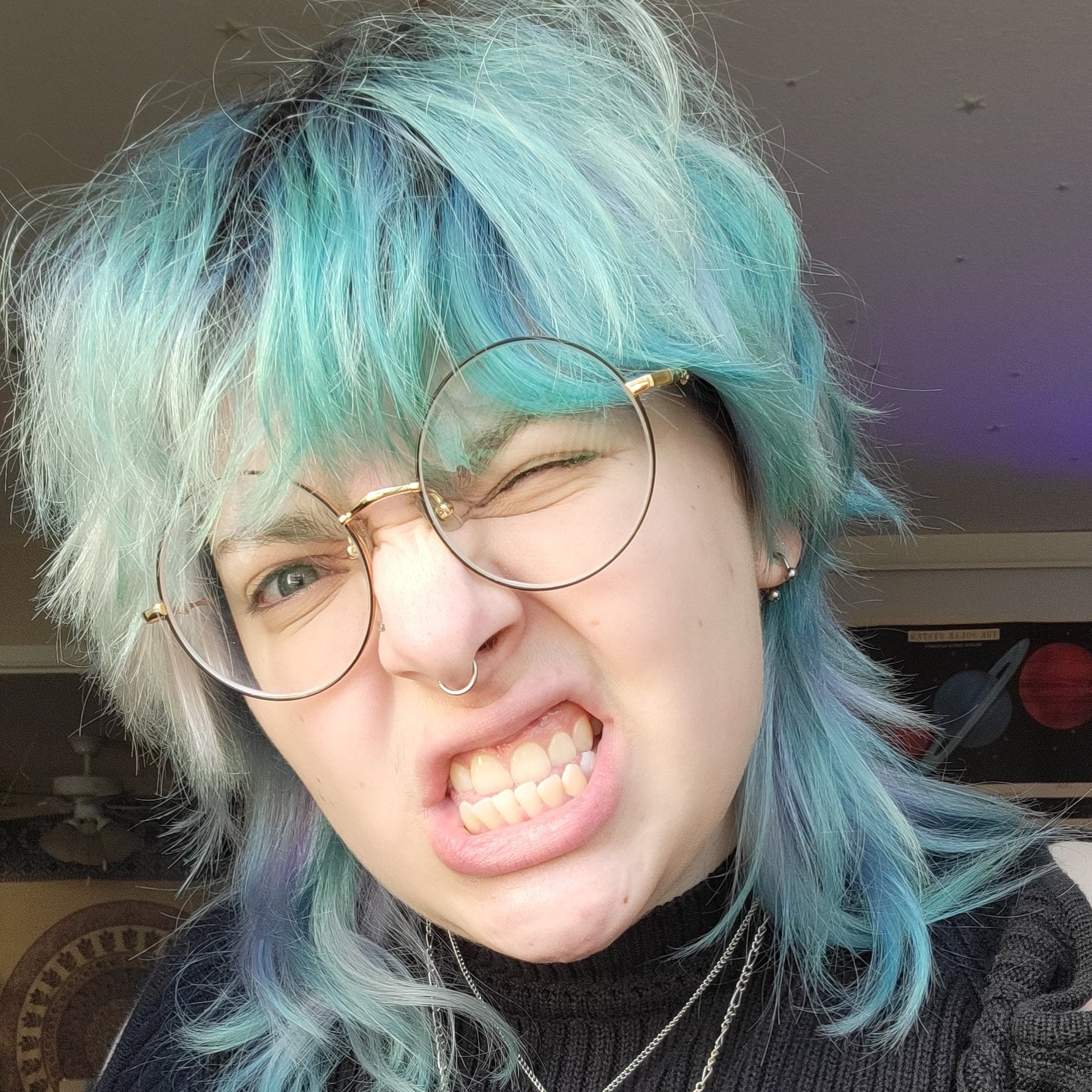 Person with teal hair wearing glasses and a dark sweater with a necklace.