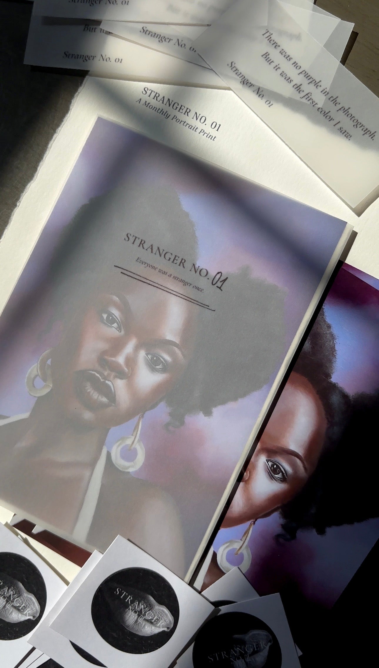 Collection of 'Stranger No. 01' art prints with stickers and vellum ephemera cards laid around it.