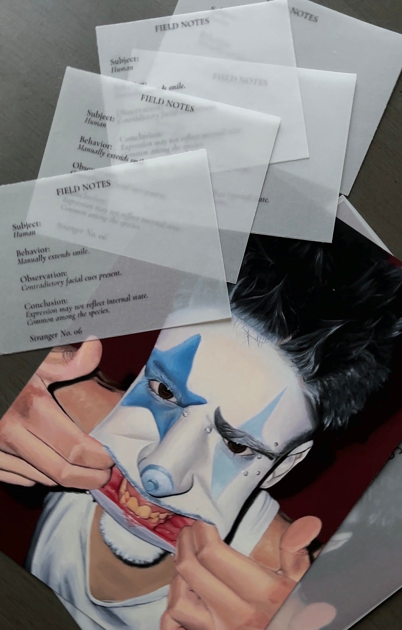 A printed art print or poster showing a stylized face with blue and white theatrical makeup, partially overlaid by several translucent labeled sheets reading FIELD NOTES, photographed on a dark surface.