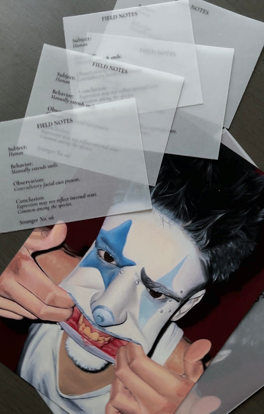 A printed art print or poster showing a stylized face with blue and white theatrical makeup, partially overlaid by several translucent labeled sheets reading FIELD NOTES, photographed on a dark surface.