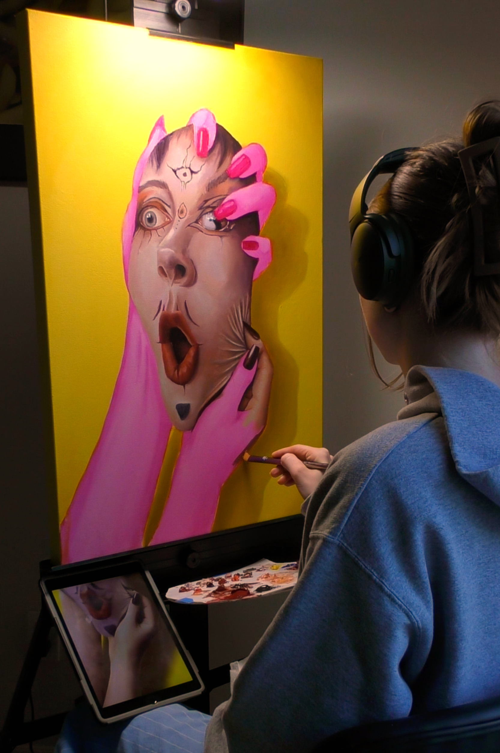 Person painting a colorful portrait of a face with pink hair on a yellow background.