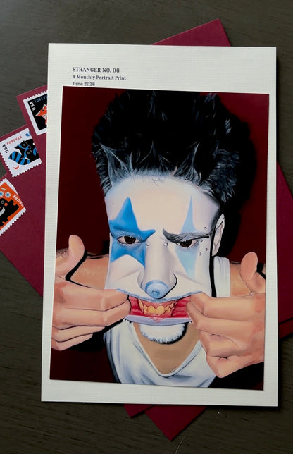 A printed portrait art piece or poster showing a stylized person with black hair, blue face paint, and hands pulling at a mouth-like structure, placed on top of a dark red envelope or backing over a dark wood surface.